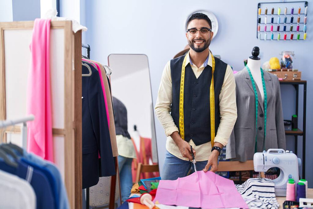 Jovem árabe homem alfaiate sorrindo pano de corte confiante na fábrica de roupas - Foto, Imagem