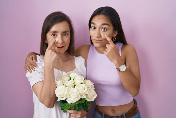 Hispanische Mutter und Tochter halten einen Strauß weißer Blumen in der Hand, die auf das Auge zeigen, das Sie beobachtet, Geste, misstrauischer Ausdruck  - Foto, Bild