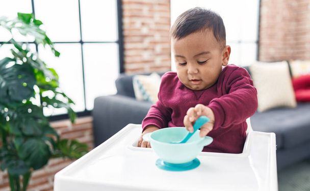 Adorable hispanic boy sitting on highchair holding spoon at home - Foto, afbeelding