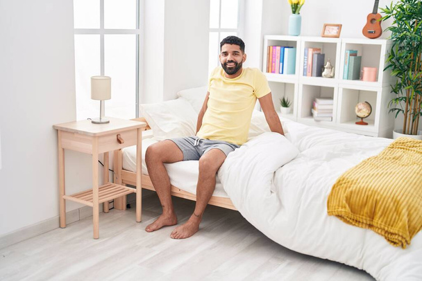 Hombre árabe joven sonriendo confiado sentado en la cama en el dormitorio - Foto, imagen