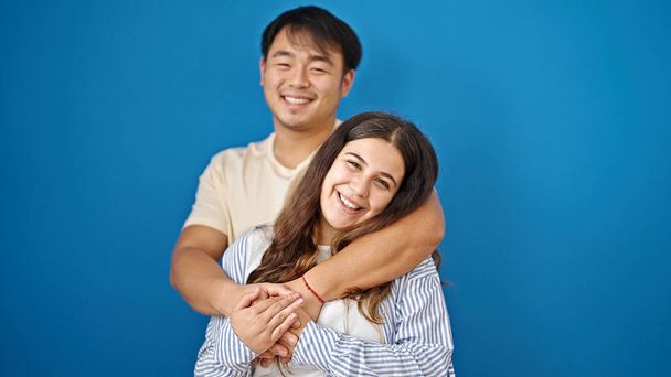 Άνδρας και γυναίκα ζευγάρι χαμογελά σίγουροι αγκαλιάζει ο ένας τον άλλον πάνω από απομονωμένο μπλε φόντο - Φωτογραφία, εικόνα