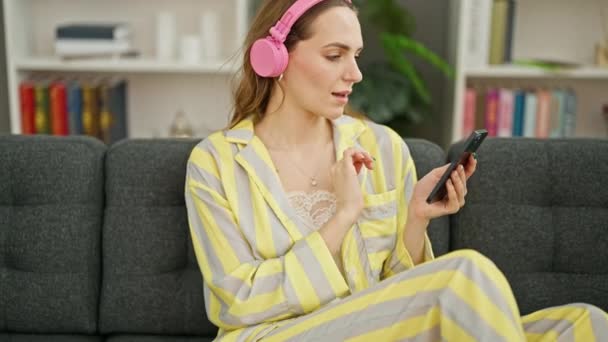 Mujer rubia joven escuchando música sentada en el sofá en casa - Metraje, vídeo
