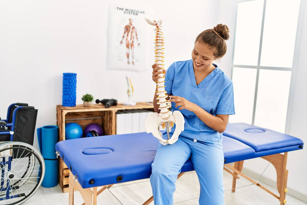Jovem bela mulher hispânica fisioterapeuta sorrindo confiante segurando modelo anatômico da coluna vertebral na clínica de reabilitação - Foto, Imagem