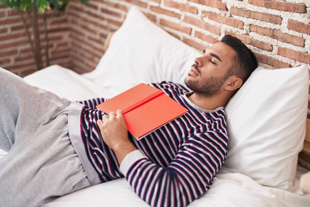 Giovane uomo ispanico che tiene libro sdraiato sul letto dormire in camera da letto - Foto, immagini