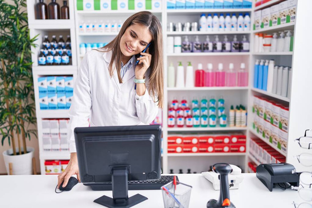 Young beautiful hispanic woman pharmacist talking on smartphone using computer at pharmacy - Photo, Image