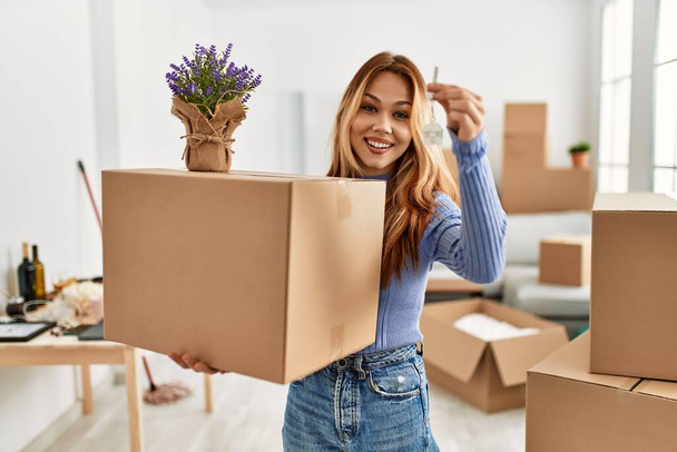 Nuori valkoihoinen nainen hymyilee luottavainen tilalla paketti laventeli kasvi ja avain uuteen kotiin - Valokuva, kuva