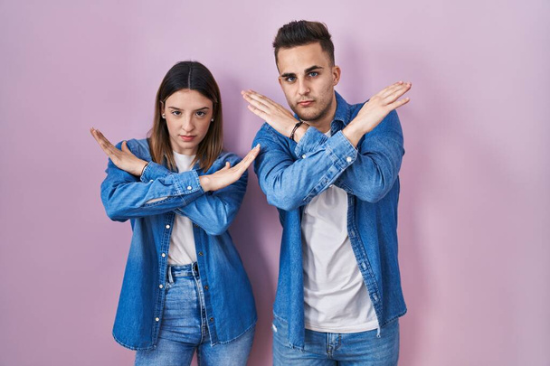 Молода пара іспаномовних, що стоять над виразом відторгнення рожевого фону, перетинає руки, роблячи негативний знак, розлючене обличчя
  - Фото, зображення