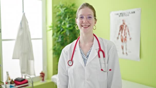 Νεαρή ξανθιά γυναίκα γιατρός χαμογελά αυτοπεποίθηση στέκεται στην κλινική - Πλάνα, βίντεο