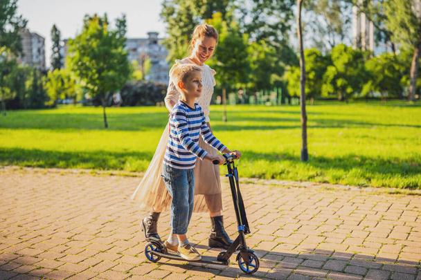 Mãe se divertindo com seu filho no parque no dia ensolarado. Rapaz está a livrar-se da scooter. - Foto, Imagem