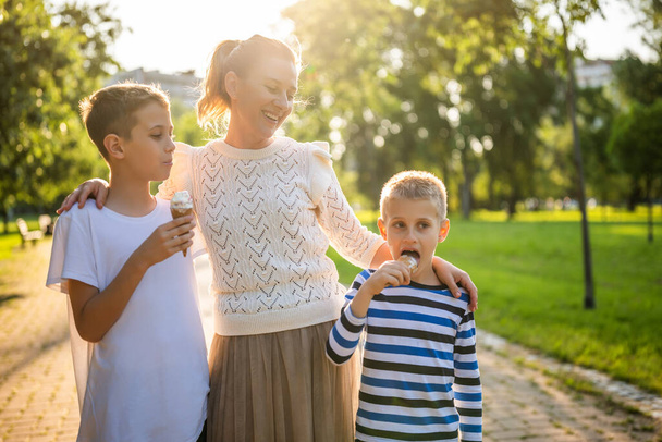 Πορτρέτο της ευτυχισμένης ανύπαντρης μητέρας με τους δύο γιους της στο πάρκο την ηλιόλουστη μέρα. Τα αγόρια τρώνε παγωτό.. - Φωτογραφία, εικόνα