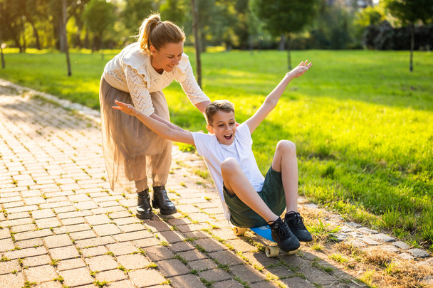 Мама веселилась с сыном в парке в солнечный день. Мальчик избавляется от скейтборда, а его мать толкает его.. - Фото, изображение