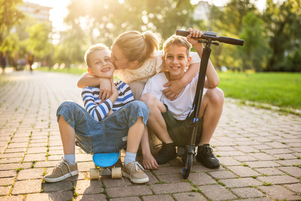 Fröhliche Mutter, die an einem sonnigen Tag mit ihren beiden Söhnen im Park Spaß hat. Sie fahren Skateboard und Roller. - Foto, Bild