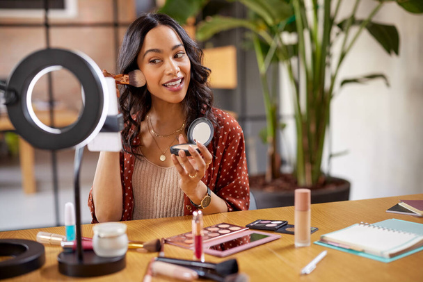 Young woman applying makeup on face with brush while recording vlog on smartphone for social media with LED ring lamp on table. Multiethnic fashion blogger creating cosmetic video. Professional hispanic beauty makeup artist vlogger livestreaming make - Foto, Imagen