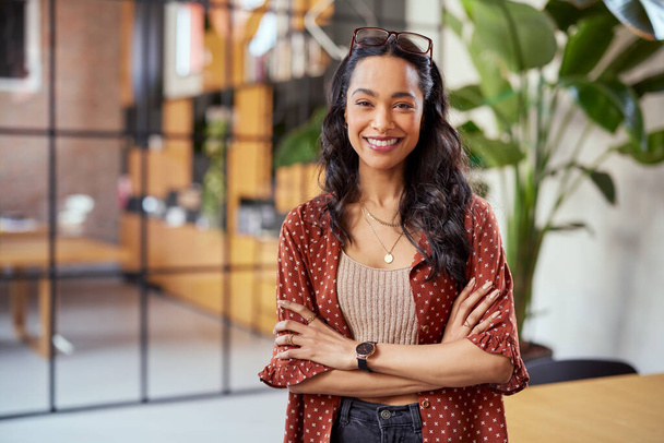 Portrait of smiling young multiethnic woman looking at camera with crossed arms. Successful latin business woman standing in modern office with copy space. Young university hispanic girl with pleasant smile and folded arms. - Zdjęcie, obraz