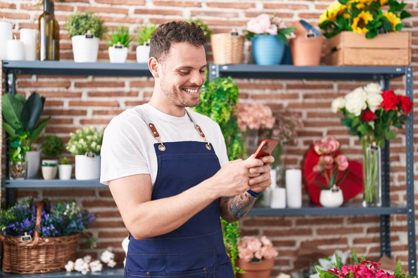 Joven florista hispano sonriendo confiado usando teléfono inteligente en floristería - Foto, imagen