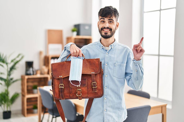 Młody Latynos z brodą pracujący w biurze trzymający walizkę i maskę bezpieczeństwa uśmiechający się z uśmiechem na dłoni i palcu.  - Zdjęcie, obraz