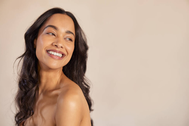 Smiling mixed race girl enjoying beauty treatment on beige background. Natural multiethnic woman looking away with copy space. Carefree latin hispanic woman with bare shoulders isolated against background looking away: skin care and bodycare concept. - Foto, Bild