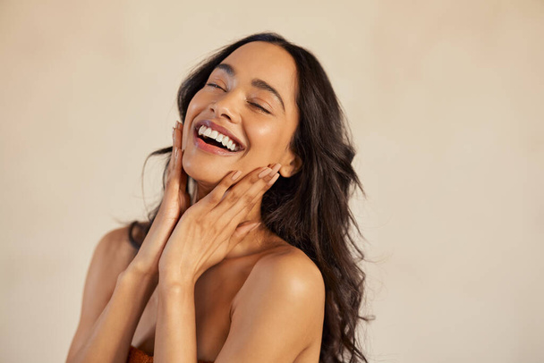 Multiethnic young woman touching her clean and healthy face against background with closed eyes. Latin hispanic woman feeling good with perfet skincare routine. Natural beauty portrait of mexican girl with black long hair. - Photo, Image