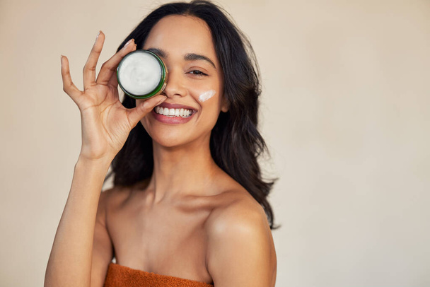 Happy attractive young woman hold green cream jar and covering eye while looking at camera with a big laugh. Portrait of multiethnic girl with bare shoulder showing natural organic cream box isolated against beige background with copy space. Mixed ra - Photo, image
