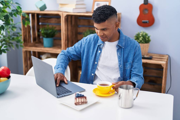 Young latin man using laptop having breakfast at home - Foto, imagen