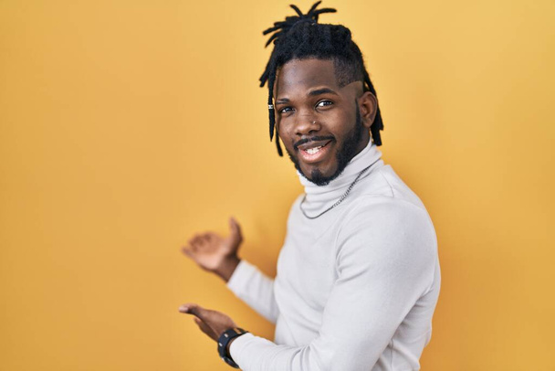 African man with dreadlocks wearing turtleneck sweater over yellow background inviting to enter smiling natural with open hand  - 写真・画像