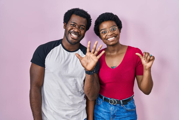 Joven pareja afroamericana de pie sobre fondo rosa mostrando y apuntando hacia arriba con los dedos número seis mientras sonríe confiado y feliz.  - Foto, imagen