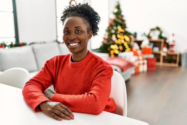 Mujer afroamericana sonriendo confiada sentada en la mesa junto al árbol de Navidad en casa - Foto, imagen