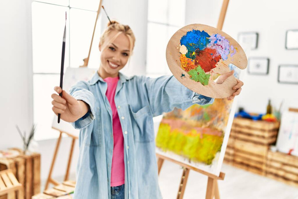 Jonge blanke vrouw kunstenaar glimlachend zelfverzekerd met penseel en palet in kunststudio - Foto, afbeelding