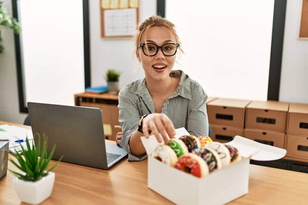 Молода біла жінка бізнесмен використовує ноутбук тримає пончик в офісі
 - Фото, зображення
