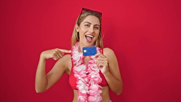 Junge blonde Touristin zeigt auf Kreditkarte und lächelt über einsamen roten Hintergrund - Foto, Bild