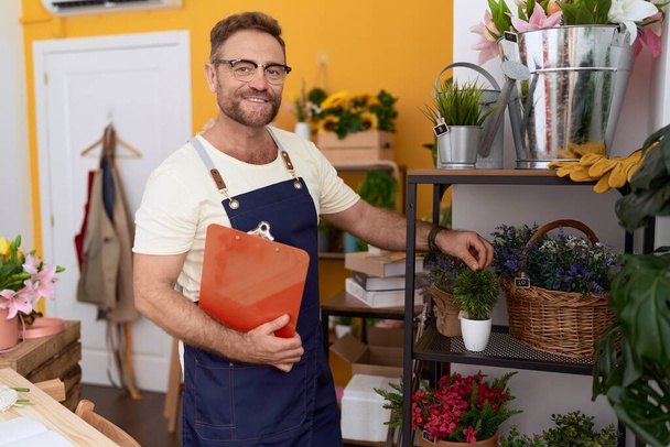 Middle age man florist touching plant holding clipboard at flower shop - 写真・画像