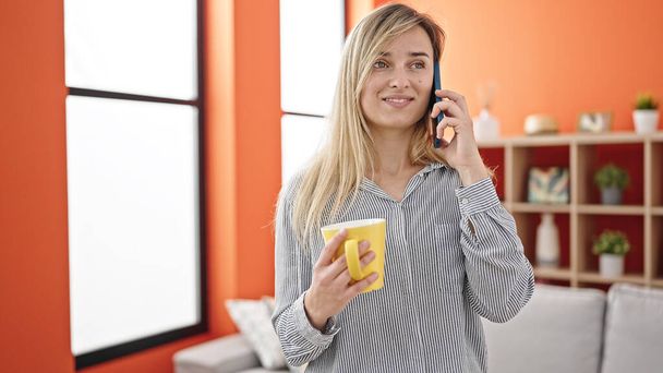 Junge blonde Frau trinkt Kaffee und telefoniert zu Hause mit dem Smartphone - Foto, Bild