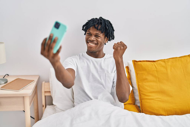 Young african man with dreadlocks taking a selfie on the bed screaming proud, celebrating victory and success very excited with raised arm  - Photo, Image
