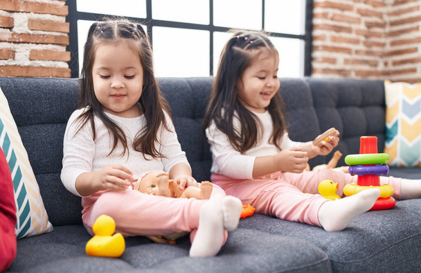 Schattige tweeling meisjes spelen met hoepels spel houden baby pop zitten op de bank thuis - Foto, afbeelding
