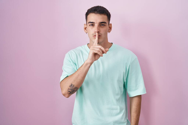 Schöner hispanischer Mann, der vor rosa Hintergrund steht und mit dem Finger auf den Lippen um Ruhe bittet. Schweigen und geheimes Konzept.  - Foto, Bild