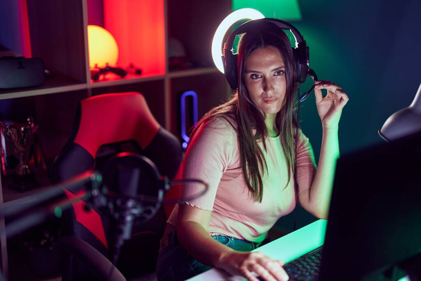 Young beautiful hispanic woman streamer sitting on table with serious expression at gaming room - Foto, Imagen