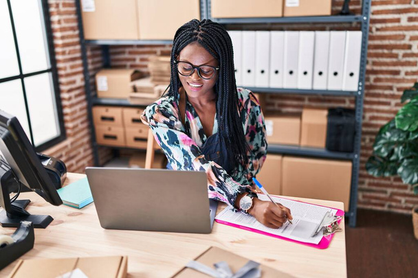 African american woman ecommerce business worker using laptop writing on document at office - Foto, Imagem