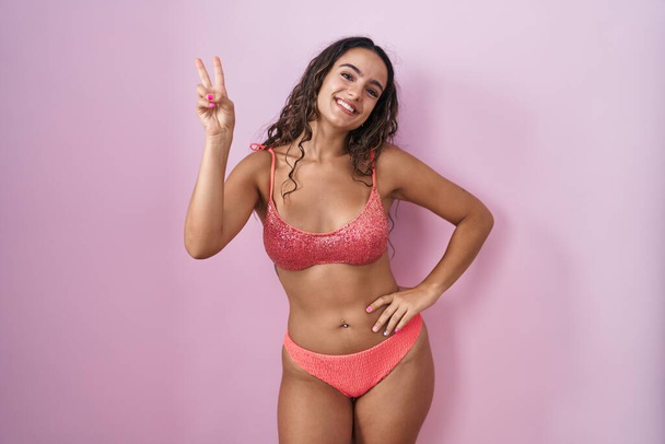 Mulher hispânica jovem vestindo lingerie sobre fundo rosa sorrindo olhando para a câmera mostrando os dedos fazendo sinal de vitória. Número dois..  - Foto, Imagem