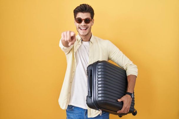 Joven hispano sosteniendo la maleta yendo de vacaciones de verano señalándote a ti y a la cámara con los dedos, sonriendo positiva y alegre  - Foto, imagen
