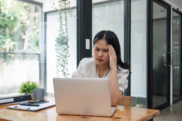Азиатские женщины напряжены во время работы на ноутбуке, усталая азиатская бизнесвумен с головной болью в офисе, чувствует себя больным на работе. - Фото, изображение