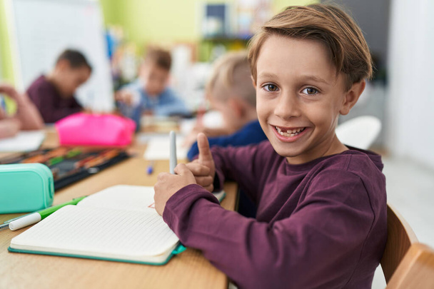 Entzückender kaukasischer Junge, der im Klassenzimmer auf ein Notizbuch schreibt und Daumen-hoch-Geste macht - Foto, Bild