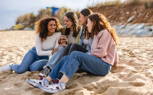 Young multiracial women laughing out loud on a sunny day - Cheerful group female friends enjoying summer vacation together on the beach sitting down- Summer vacation - Photo, Image