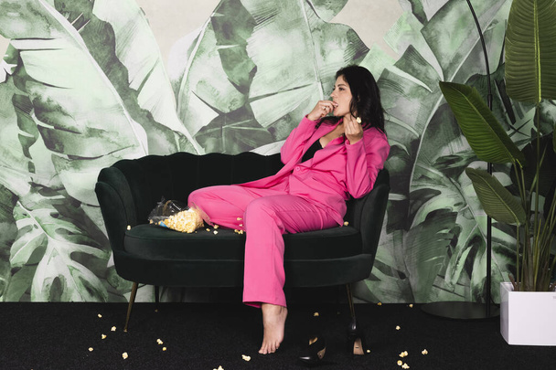 Молодая женщина в розовом костюме проводит время одна дома, ест попкорн на диване и смотрит телевизор. грязная комната с разбросанным попкорном - Фото, изображение