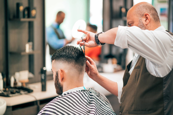Vue latérale du coiffeur mature en chemise et gilet coupant les cheveux foncés du client masculin en cape rayée avec ciseaux sur fond flou - Photo, image