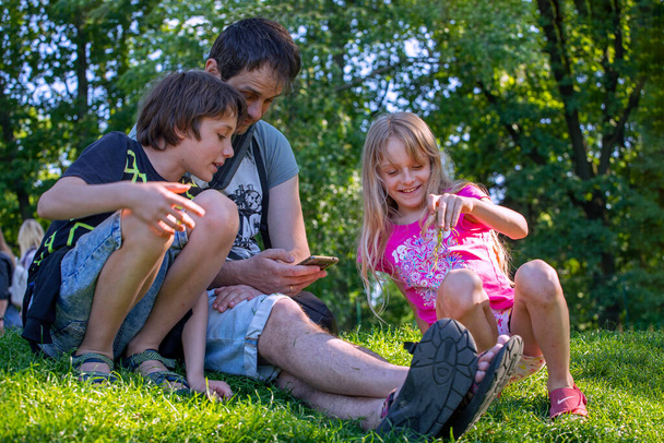 glückliche Vater mit Sohn und Tochter betrachten die Gadget-Informationen, während sie auf dem Gras in der Natur sitzen. Gemeinsamer Familienurlaub. Zwei Generationen Vater und Sohn und Tochter - Foto, Bild