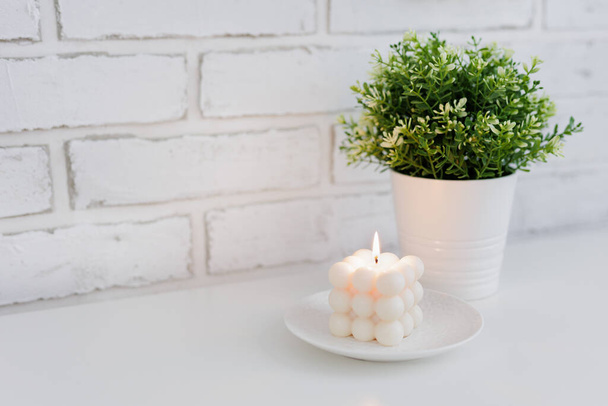 Burning white bauble candle with green plant on the table near brick wall - 写真・画像