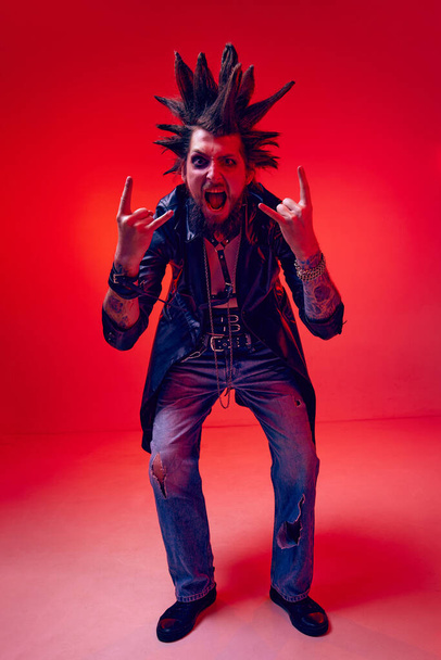 Rock and roll. Emotive, expressive young man, punk with makeup and strange hairdo posing over red studio background in neon light. Concept of music, lifestyle, subculture, art, youth, human emotions - Photo, image