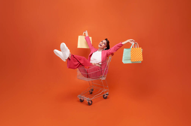 幸せな笑顔トロリーとショッピングバッグを持つファッショナブルな女性は、オレンジの背景に隔離されたショッピングプロモーション夏の販売を歩く. - 写真・画像