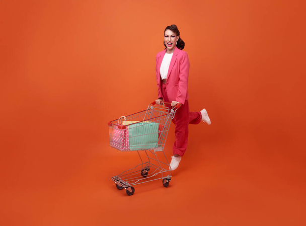 heureux souriant femme à la mode avec chariot et sacs à provisions marche shopping promotion été vente isolé sur fond orange. - Photo, image