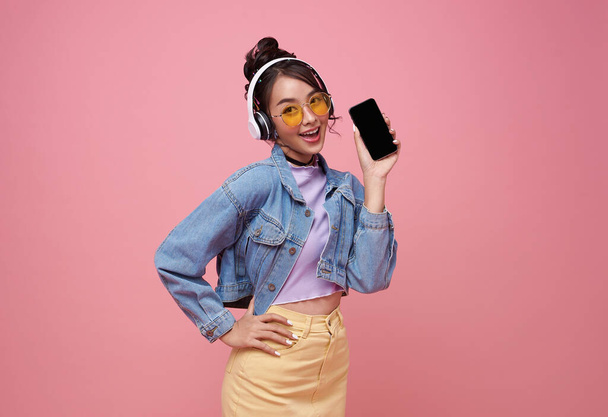 jong aziatische tiener vrouw tonen smart phone ze luisteren muziek in hoofdtelefoon geïsoleerd op roze achtergrond. - Foto, afbeelding
